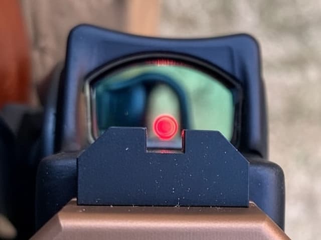 Vista a través de una mira de punto rojo montada en una pistola Glock, configurada con sistema co-witness para adquisición inmediata de blancos.